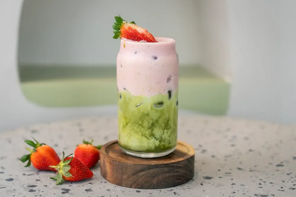 Green matcha base under a thick pink cream layer, a halved strawberry on the rim and three whole berries beside the glass