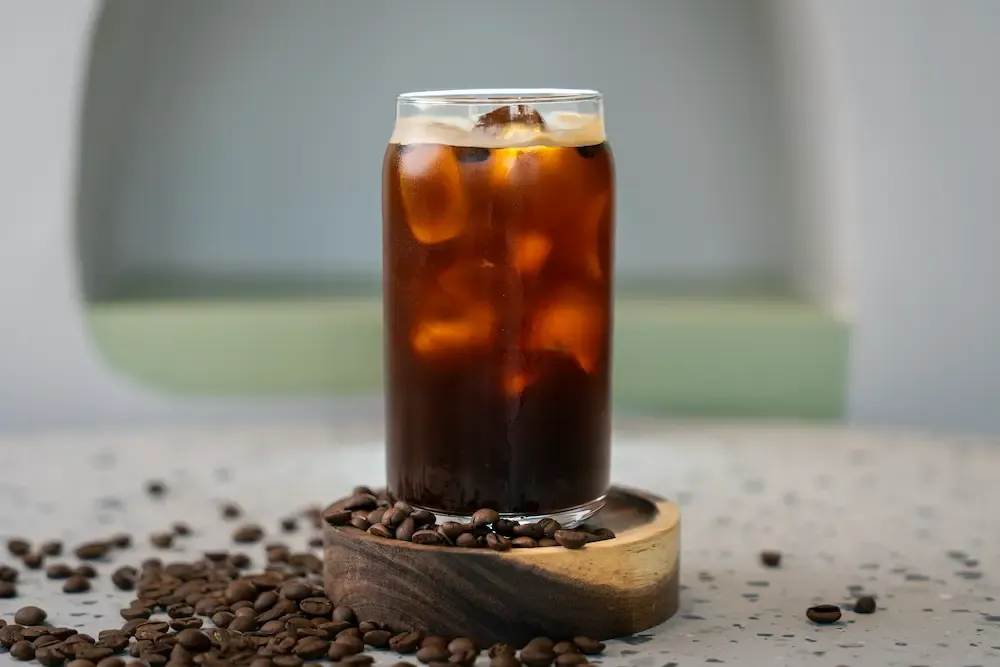 Dark iced coffee in a can-shaped glass, pale crema on top and ice cubes glowing amber, roasted beans strewn around the base