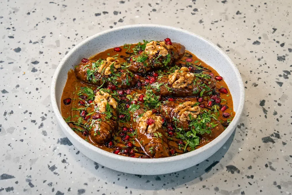 Six kibbeh torpedoes sitting in a rust-red sauce, each capped with a walnut half and strewn with chopped parsley and pomegranate seeds