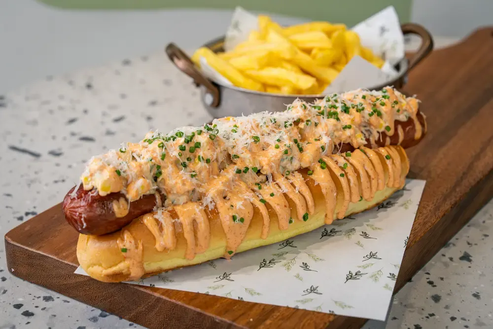 Long sausage in a split roll under creamy sweetcorn relish, grated cheese and chopped chives, orange mayo zigzagged along the bun