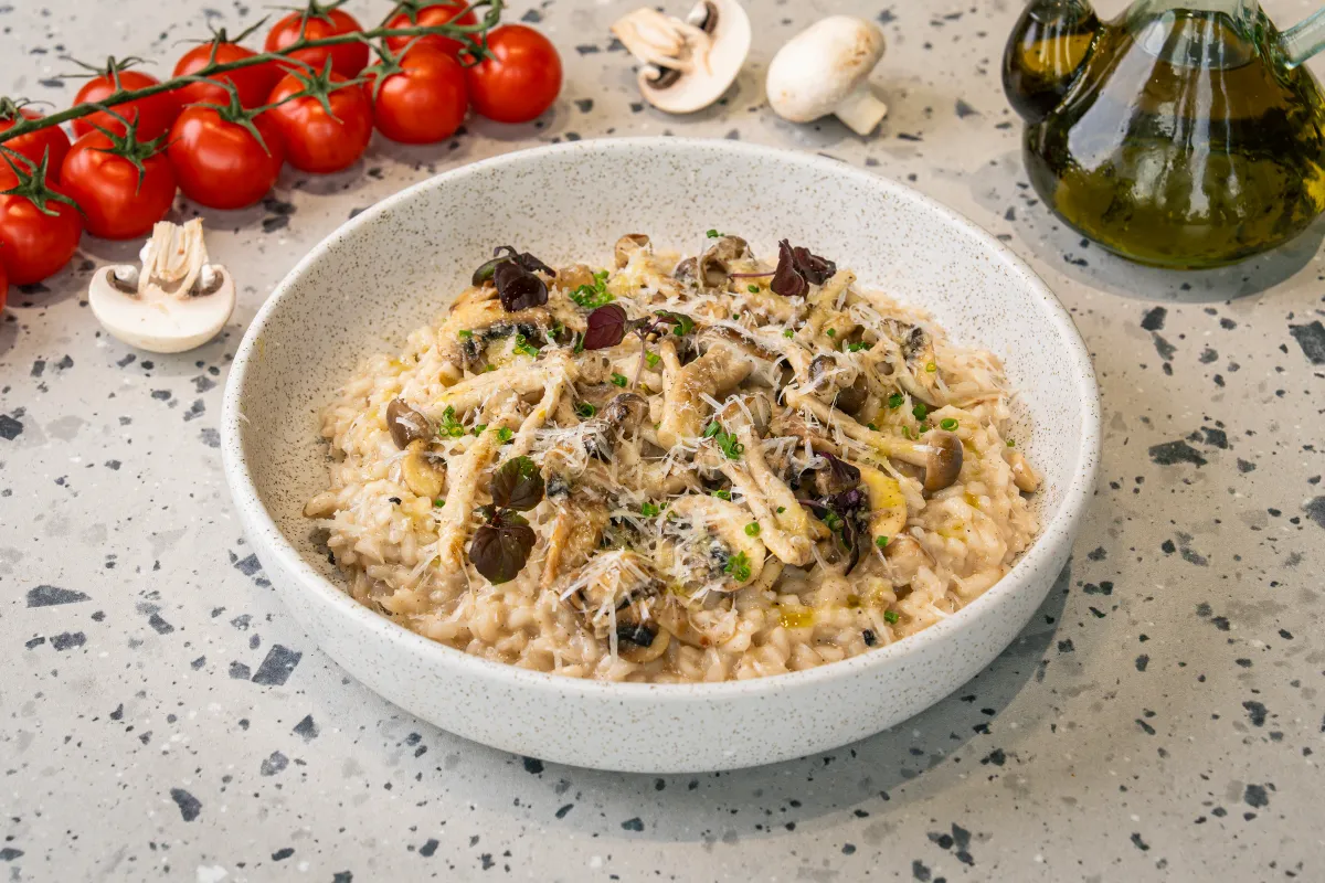 Pale creamy risotto in a speckled white bowl under torn mushrooms, grated parmesan, snipped chives and small purple basil leaves