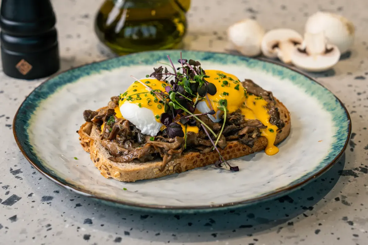 Sourdough heaped with dark sautéed mushrooms and poached eggs under yellow hollandaise running off the edge, topped with purple microherbs