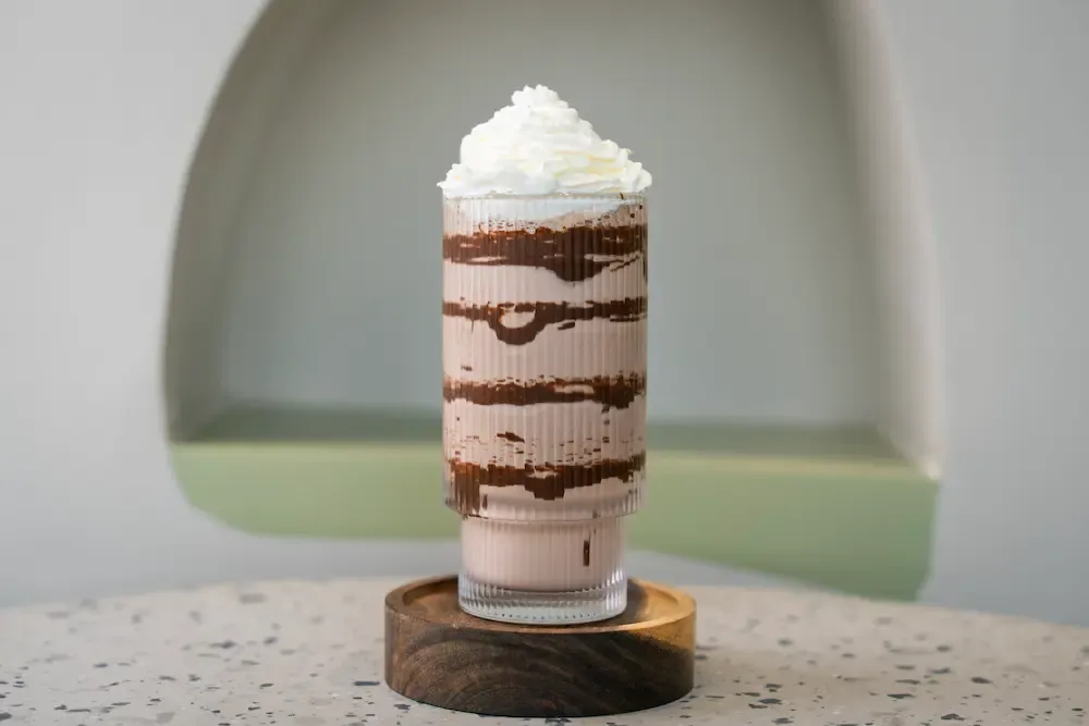 Ribbed glass of pale brown shake streaked with chocolate sauce down the inside, under a tall swirl of whipped cream
