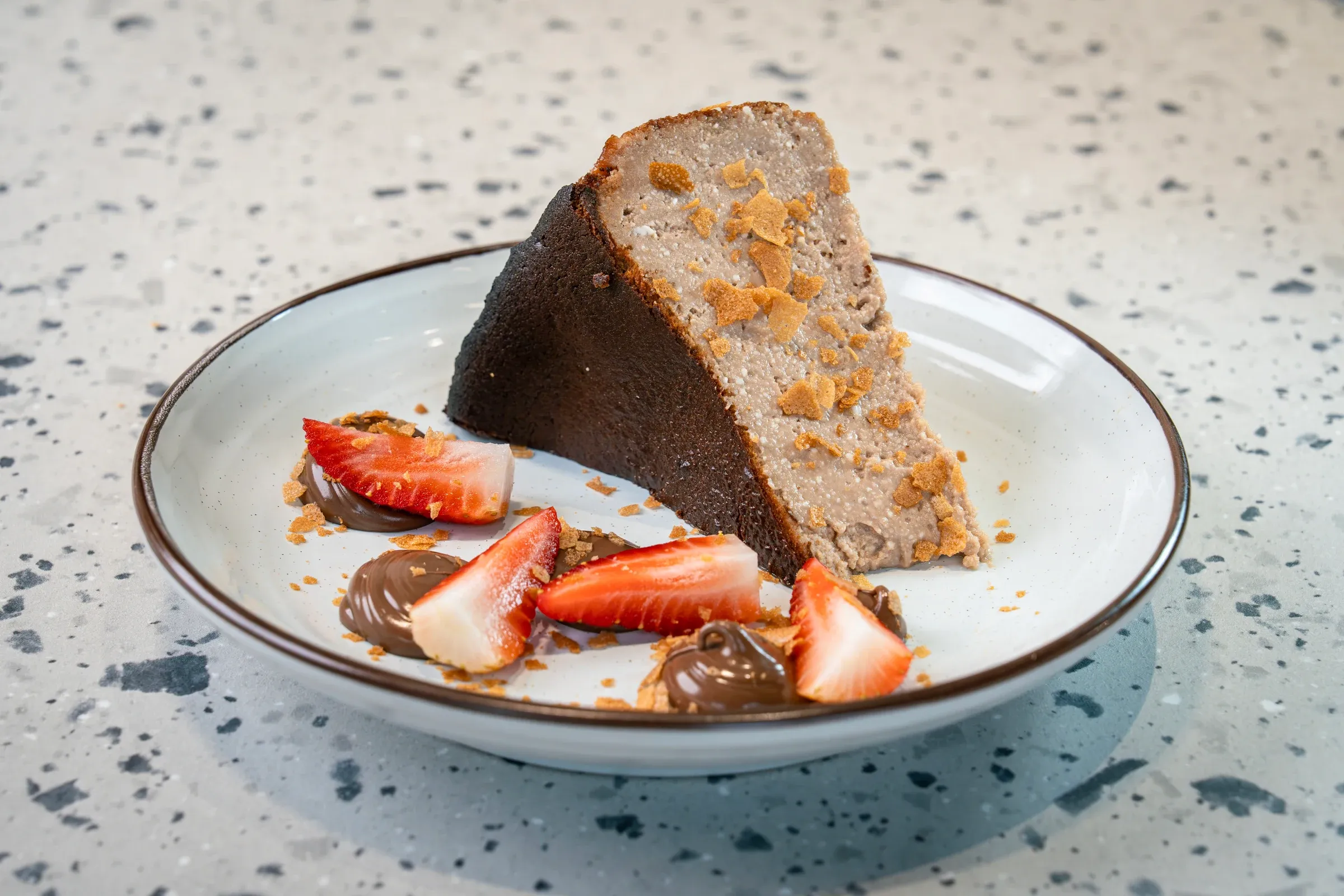 Thick wedge of pale coffee-coloured cheesecake with a dark scorched crust, biscuit crumbs, sliced strawberries and chocolate dollops