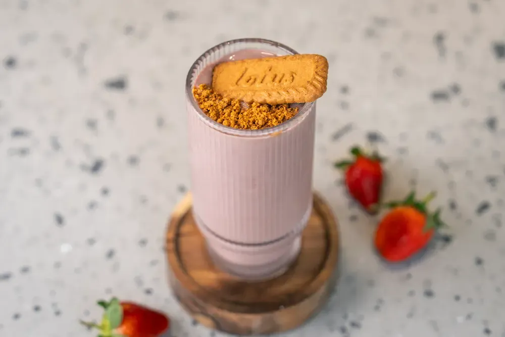 Pale pink shake in a ribbed glass topped with a Lotus biscuit and spiced crumbs, strawberries scattered around the wooden coaster
