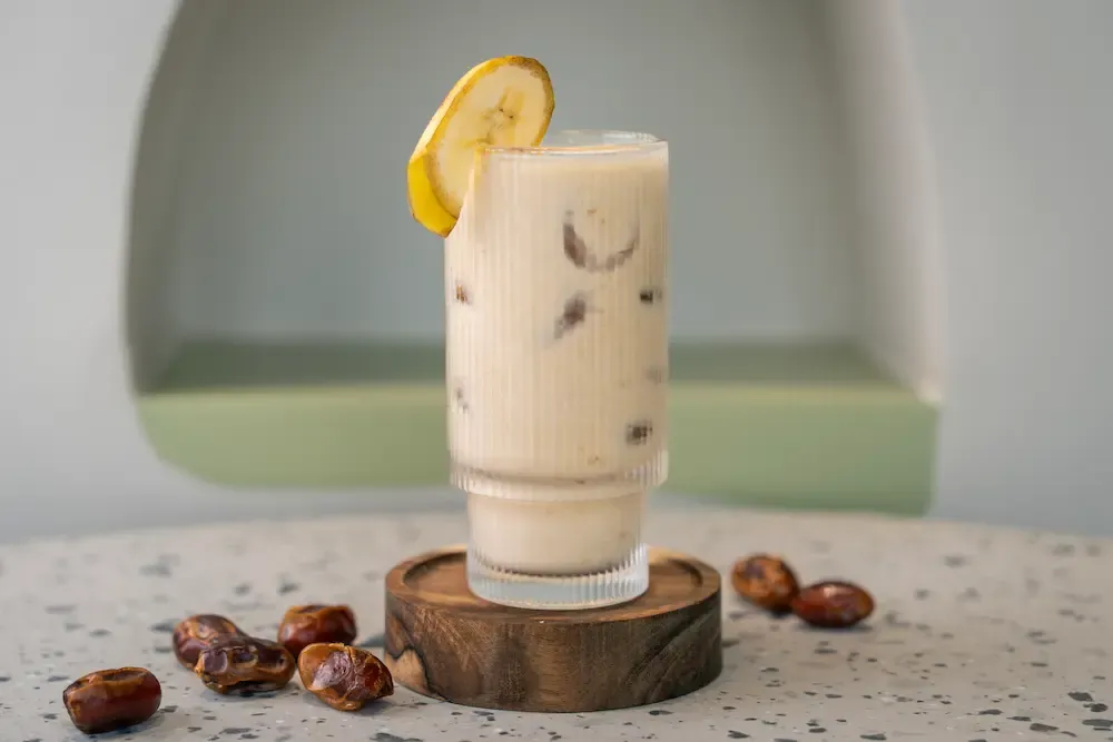 Cream-coloured shake flecked with chopped dates in a ribbed glass, a dried banana slice on the rim and whole dates scattered nearby