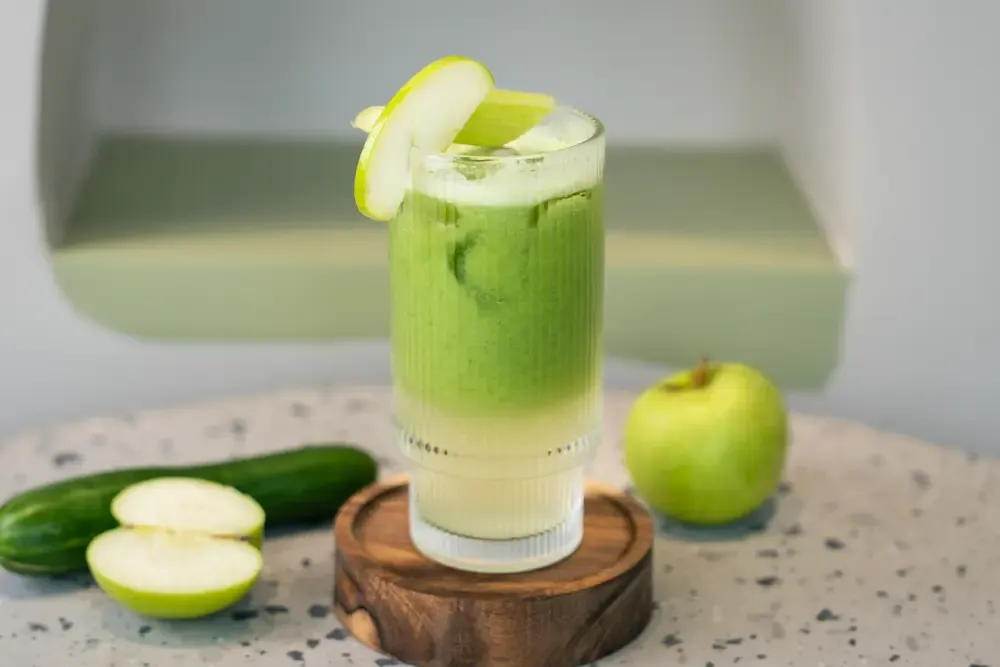 Layered green juice in a ribbed glass with an apple slice and celery baton at the rim; a cucumber and cut apples alongside
