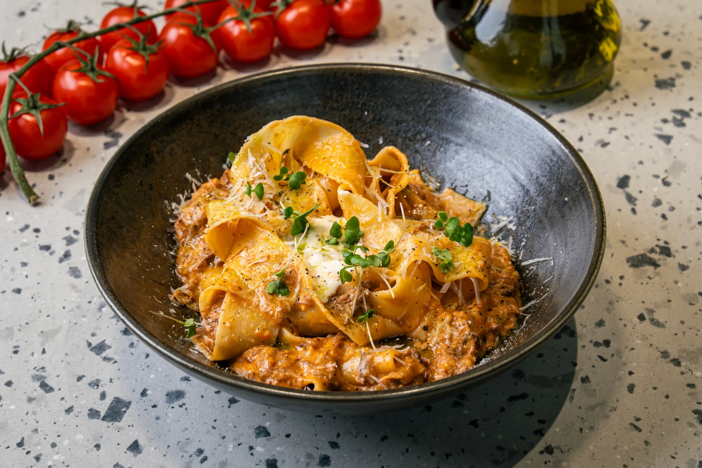 Wide pappardelle ribbons folded over a rust-red rib ragout in a matte black bowl, with a spoon of cream, micro herbs and shaved parmesan
