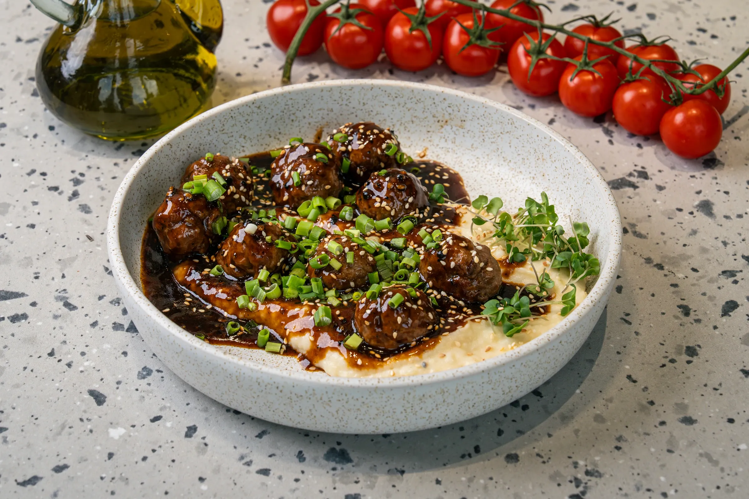 Glossy dark-glazed meatballs crowded over mashed potato, scattered with sesame seeds and sliced spring onion, cress at the bowl's edge