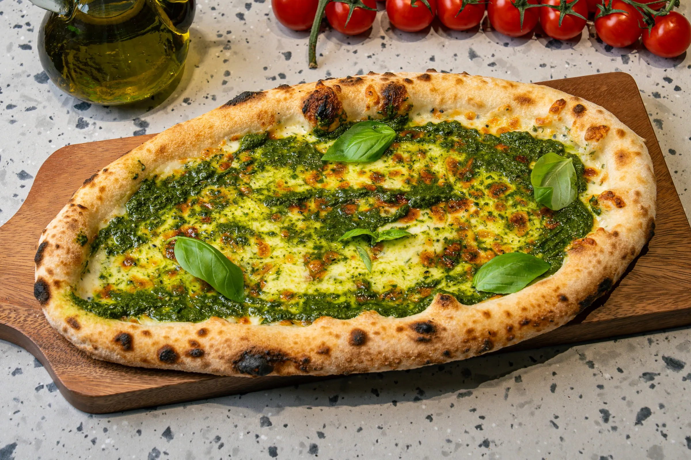 Oval flatbread with a puffed, blistered crust, swirls of green pesto over melted cheese and fresh basil leaves, on a wooden board