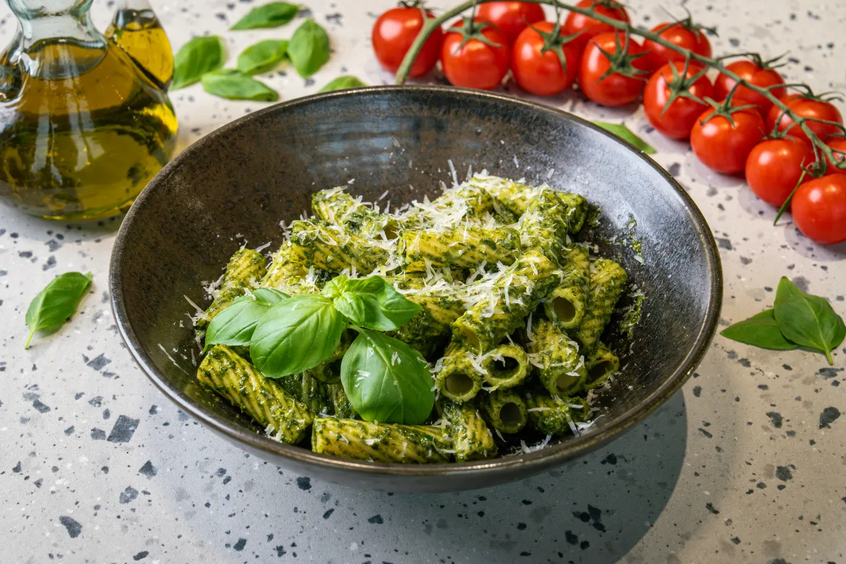 Rigatoni tubes coated in thick green pesto in a dark stoneware bowl, under grated parmesan with two fresh basil leaves on top