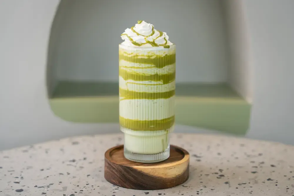 Pale green shake banded with darker pistachio sauce inside a ribbed glass, whipped cream on top drizzled with more green sauce