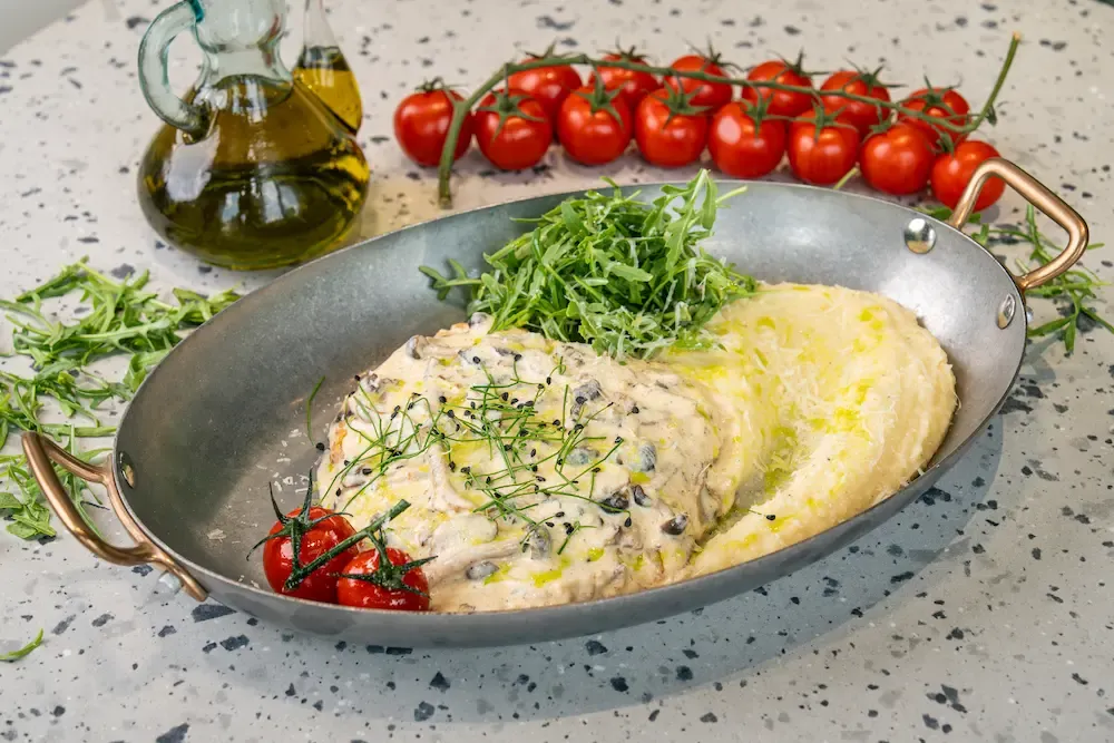 Breaded chicken under pale mushroom cream sauce in a copper-handled metal pan, beside mash, a heap of rocket and vine cherry tomatoes