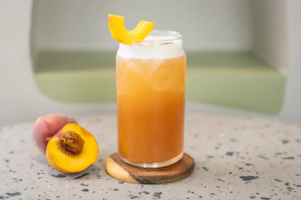 Cloudy peach-amber tea over ice in a can-shaped glass, a peach slice notched over the rim, halved peach with its stone alongside