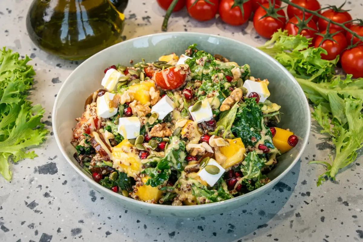 Sage-green bowl heaped with red and white quinoa, curly kale, feta cubes, mango, pomegranate seeds and walnuts under a yellow dressing