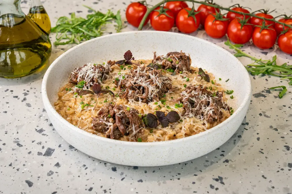Shredded dark braised beef piled over pale risotto in a speckled bowl, under a heavy dusting of grated parmesan, chives and purple basil