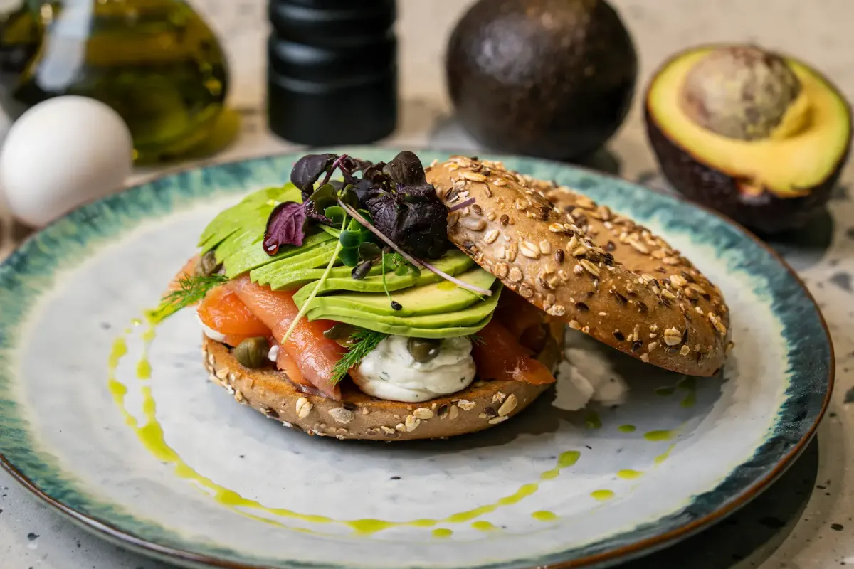 Seeded bagel open on a teal-rimmed plate, layered with cream cheese, smoked salmon, fanned avocado and capers, the lid leaning alongside