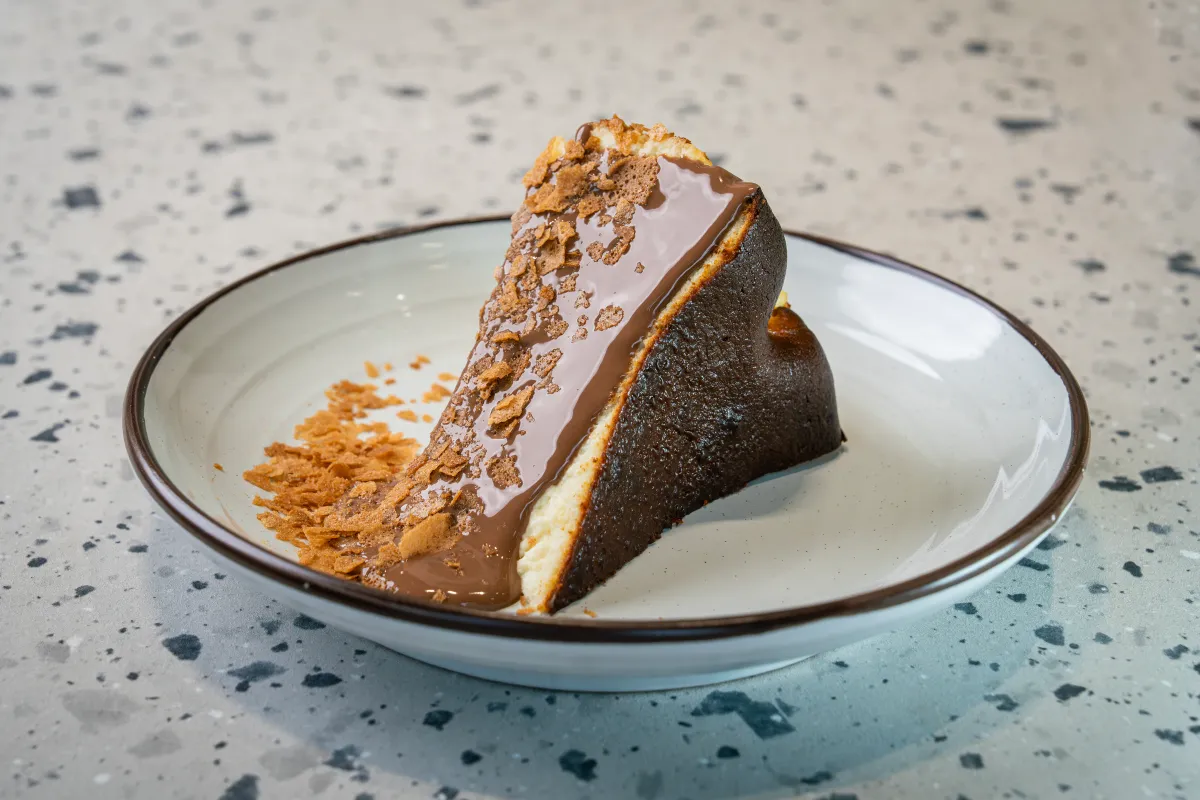 Slice tipped on its side, cut face under a thick chocolate glaze and biscuit crumb, burnt-dark crust behind, on a brown-rimmed plate