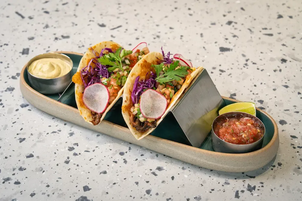 Two soft tacos of shredded beef topped with red cabbage, coriander and radish, on a long platter with pots of guacamole and salsa