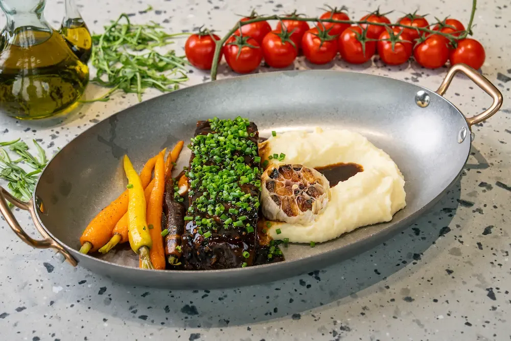 Dark glazed rib coated in chopped chives in an oval steel pan, with baby carrots, a roasted garlic bulb and a swirl of mashed potato