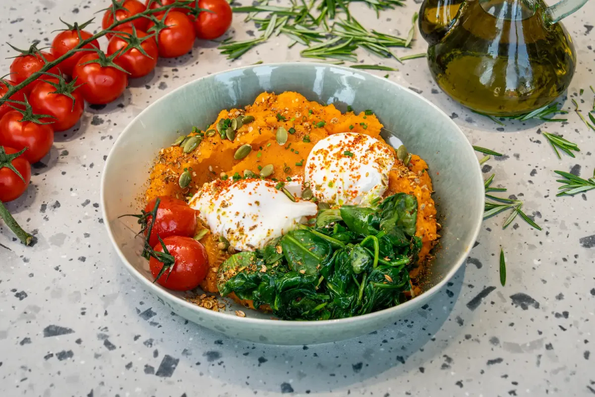 Mashed orange sweet potato in a pale green bowl with two poached eggs dusted red, wilted spinach, vine tomatoes and pumpkin seeds
