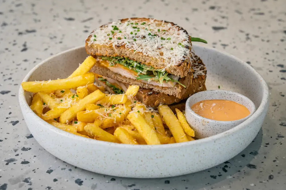 Toasted sourdough sandwich under a blanket of grated cheese and chives, chicken and rocket inside, beside fries and a pink dip