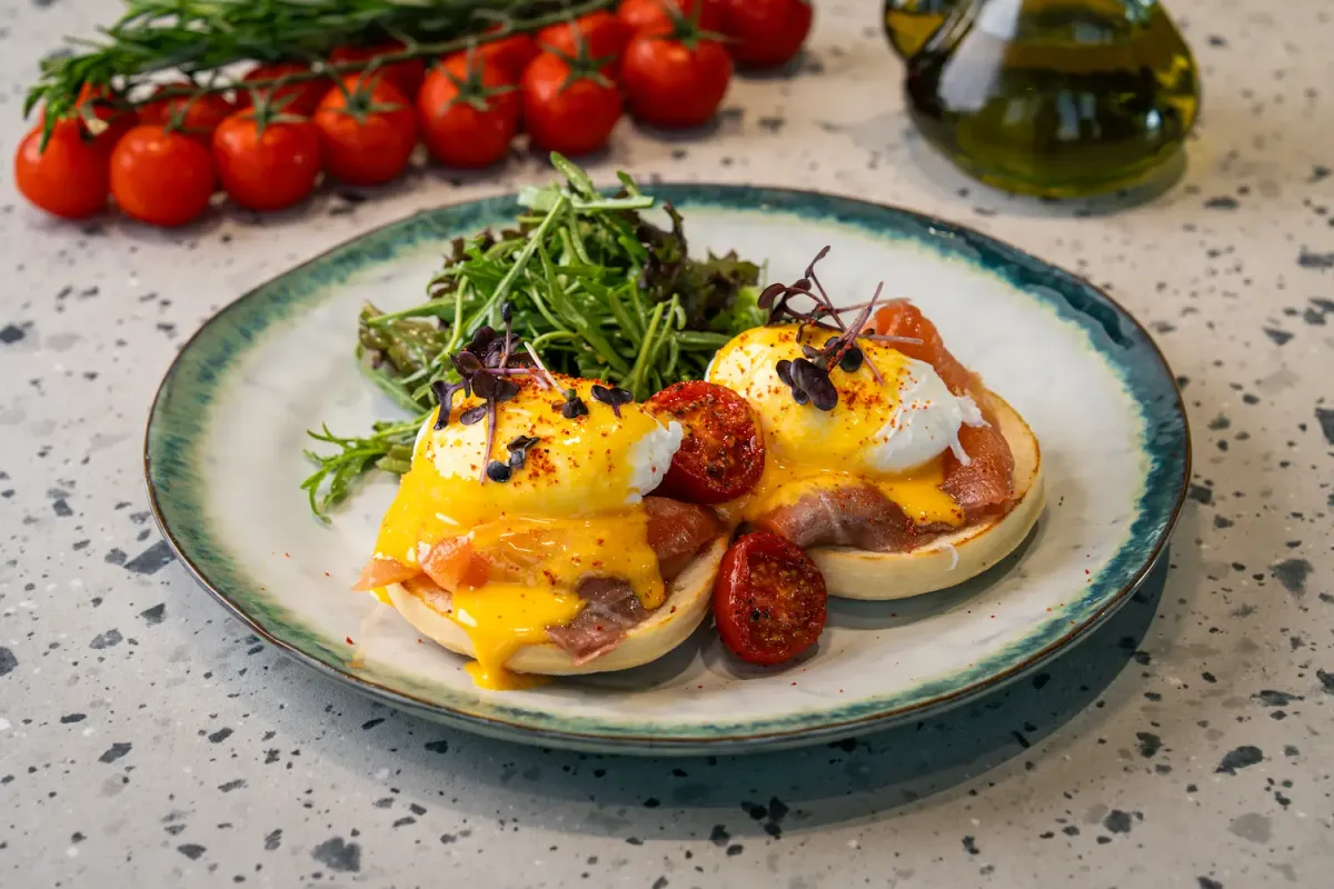 Two muffin halves under glossy yellow hollandaise, poached eggs cut open and running, with pink smoked salmon, roast tomato and rocket