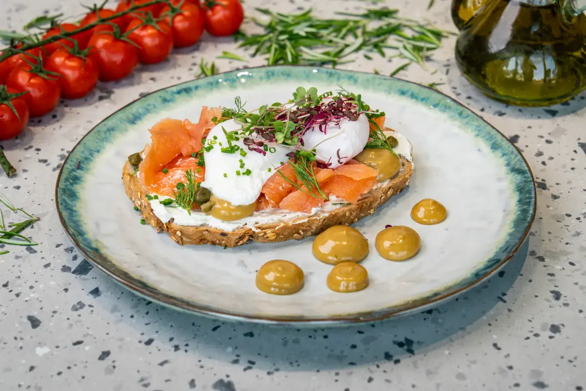 Thick sourdough spread with cream cheese under folded salmon, poached eggs and capers, with dots of mustard beside it on the plate