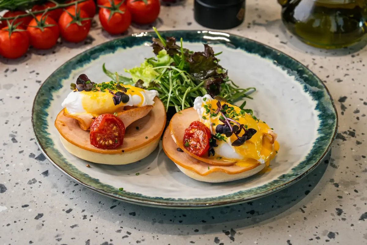 Two muffin halves topped with folded turkey slices, roast tomato and poached eggs under yellow hollandaise, salad leaves piled behind