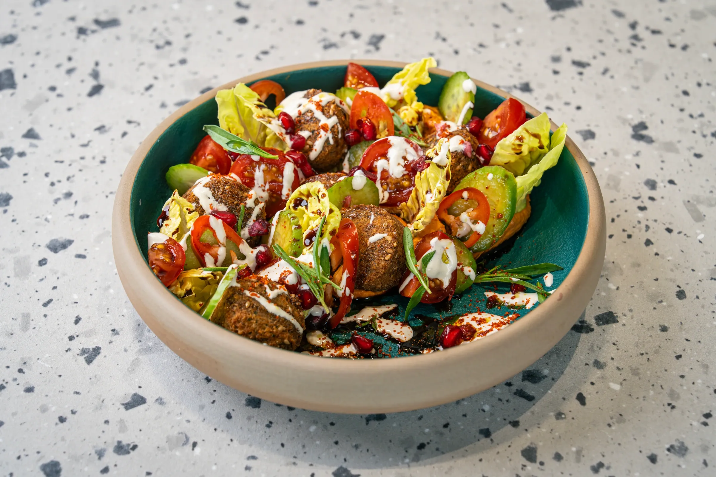 Craggy falafel balls piled over baby gem, tomato and cucumber in a deep teal bowl, laced with white tahini and pomegranate seeds