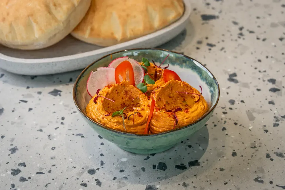 Two mounds of orange-tinted hummus dusted with paprika, topped with radish, tomato and red micro leaves; pitta on a tray behind