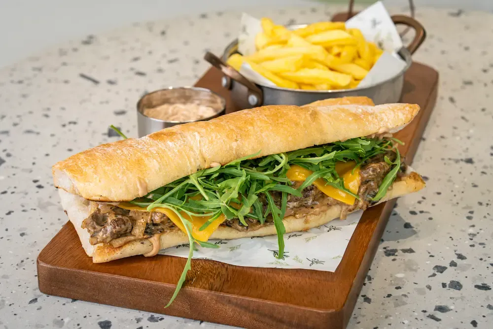 Split baguette filled with creamy mushrooms, cheddar and rocket on a wooden board, with a metal pot of chips and pink dipping sauce