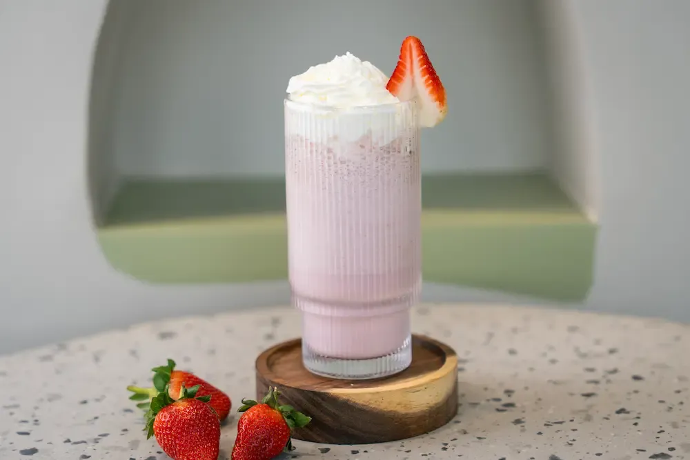 Pale pink shake in a ribbed glass with whipped cream and a halved strawberry on the rim, three whole berries at the base