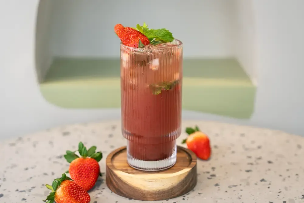 Deep pink drink over ice in a tall fluted glass on a wooden coaster, mint sprig and strawberry slice on the rim, whole berries alongside