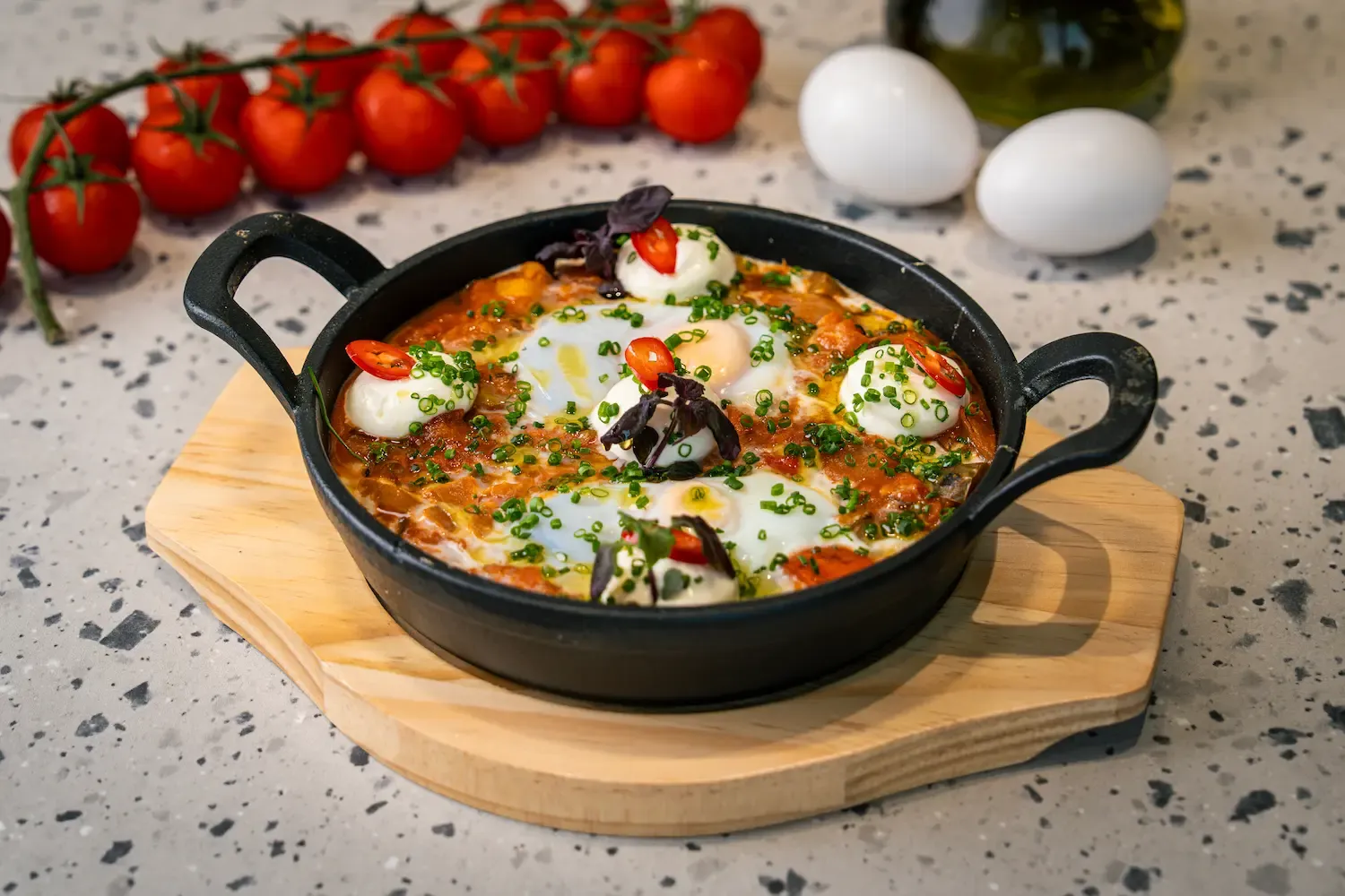Tomato and pepper stew with four soft-set eggs in a small cast-iron pan on a wooden board, scattered with chives, chilli and purple basil