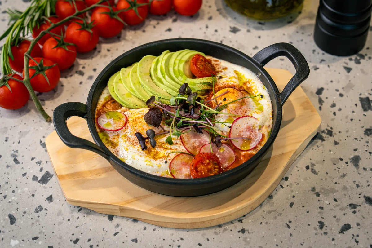 Cast-iron pan of eggs in red sauce, half covered by fanned avocado, with radish slices, paprika, roast tomato and micro herbs, on a board