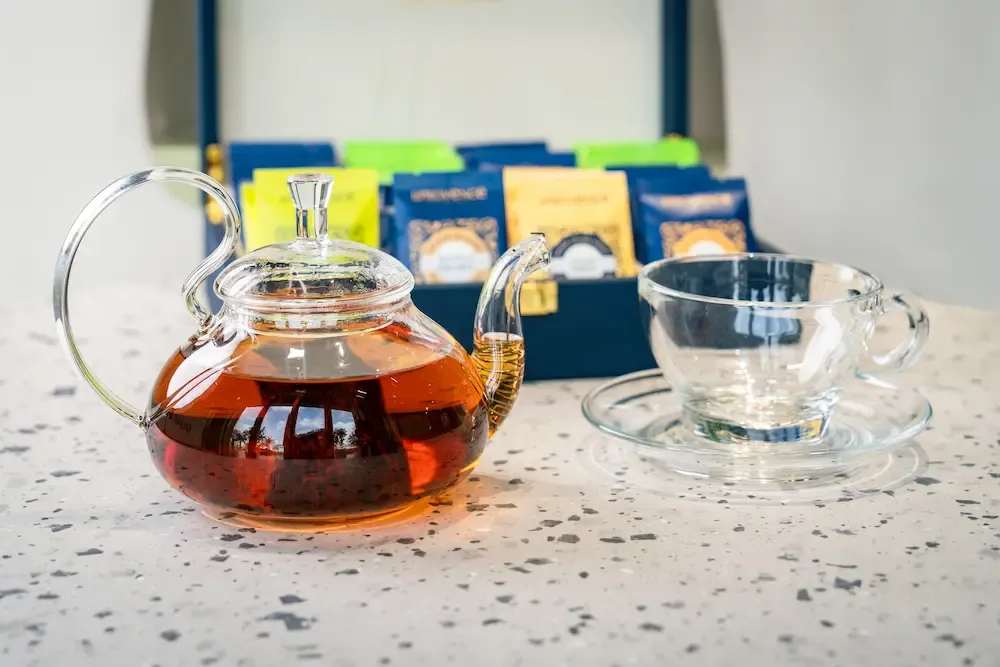 Glass teapot of amber tea with leaves settled at the base, empty glass cup and saucer beside it, navy box of teabags behind