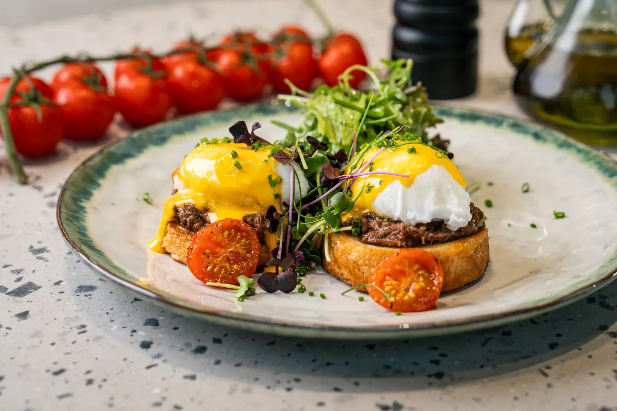 Two toast slices piled with shredded braised short rib and poached eggs under yellow hollandaise, roast tomato halves and micro leaves