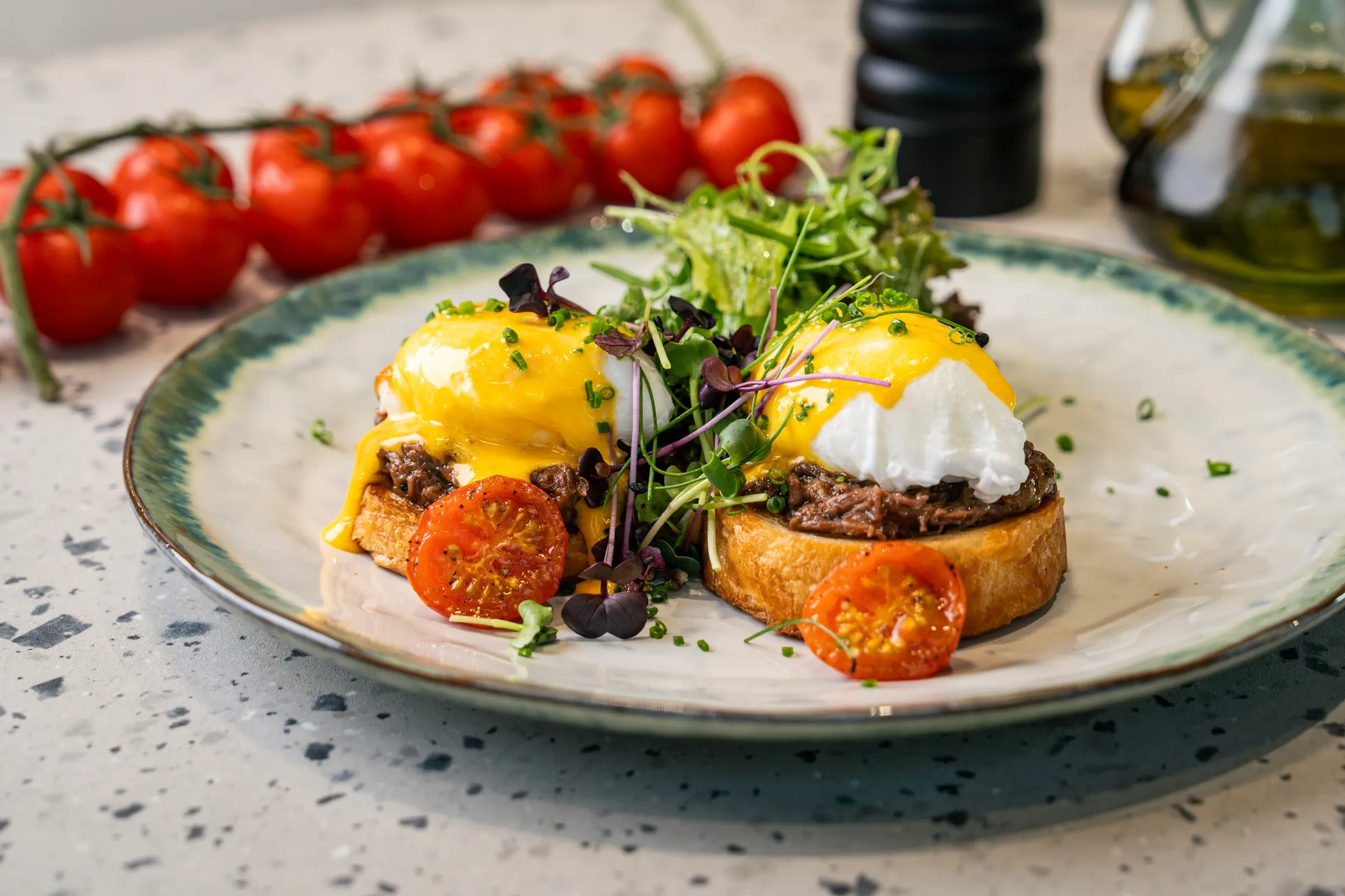 Two toast slices piled with shredded braised short rib and poached eggs under yellow hollandaise, roast tomato halves and micro leaves