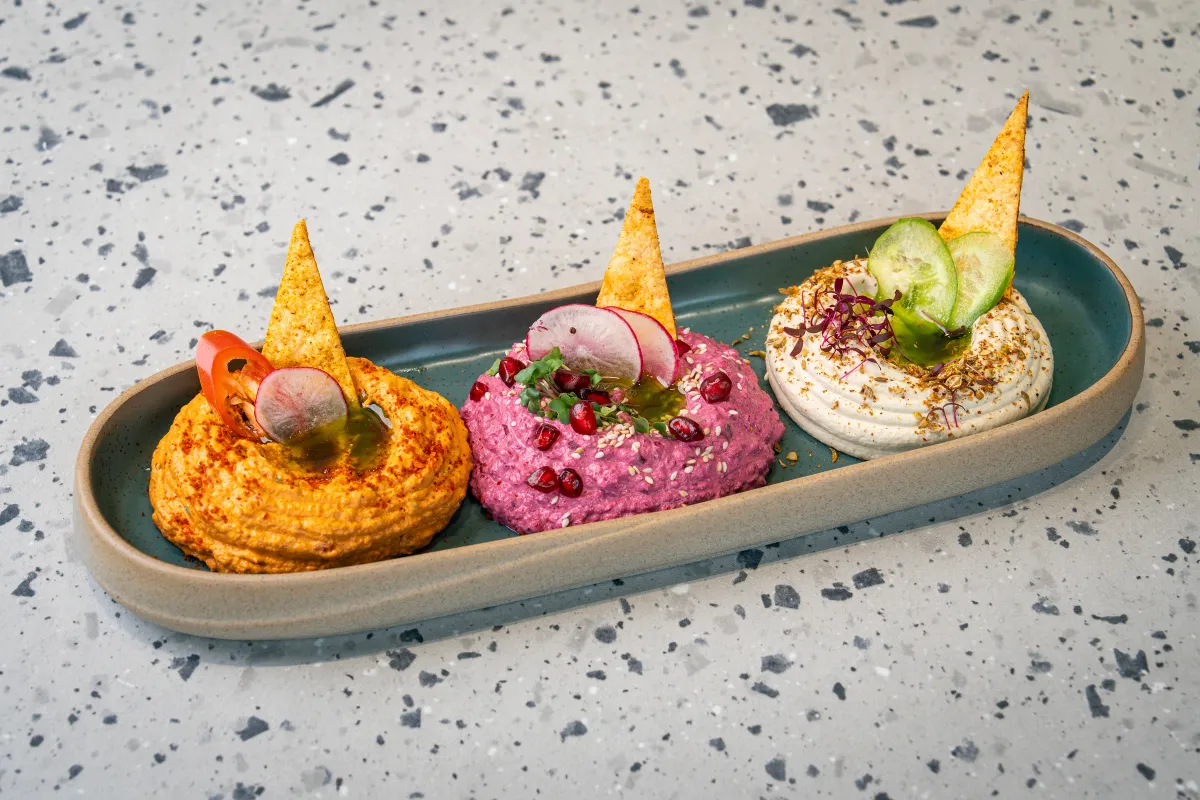 Three dips lined up in a long oval dish - paprika-orange, magenta beetroot, and white with za'atar - each spiked with a tortilla wedge