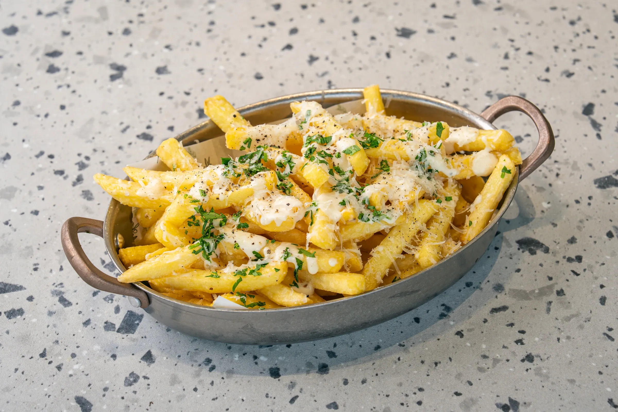 Pale golden fries heaped in a two-handled metal pan under melted cheese, grated parmesan, cracked pepper and chopped parsley
