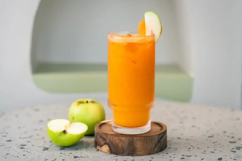 Vivid orange carrot juice over ice in a ribbed glass, a slice of green apple on the rim and whole green apples beside the coaster