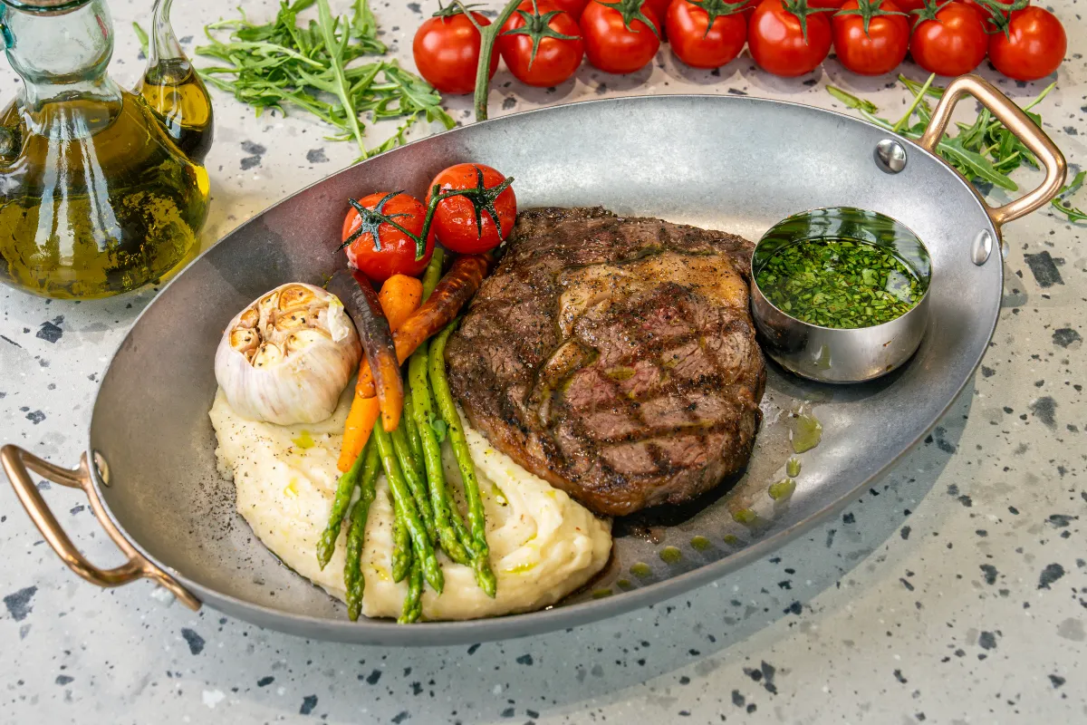 Thick grilled rib eye with crosshatch char marks in a steel pan, with mash, asparagus, baby carrots, roasted garlic and chimichurri pot