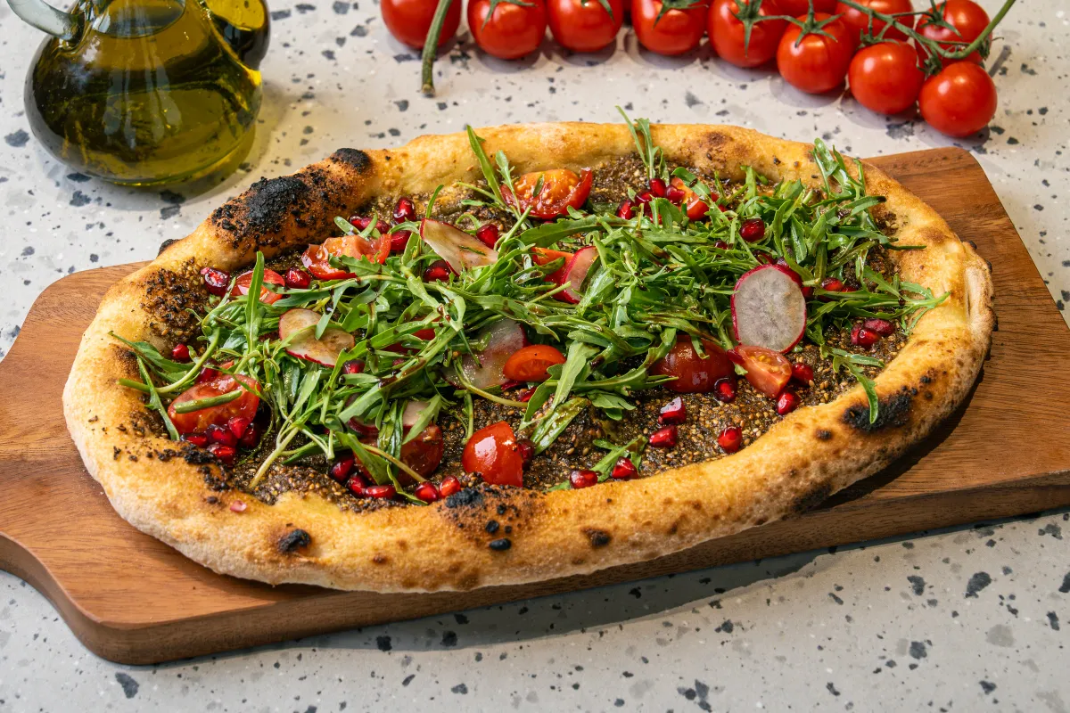 Oval flatbread spread with dark green za'atar, piled with rocket, halved cherry tomatoes, radish slices and pomegranate seeds