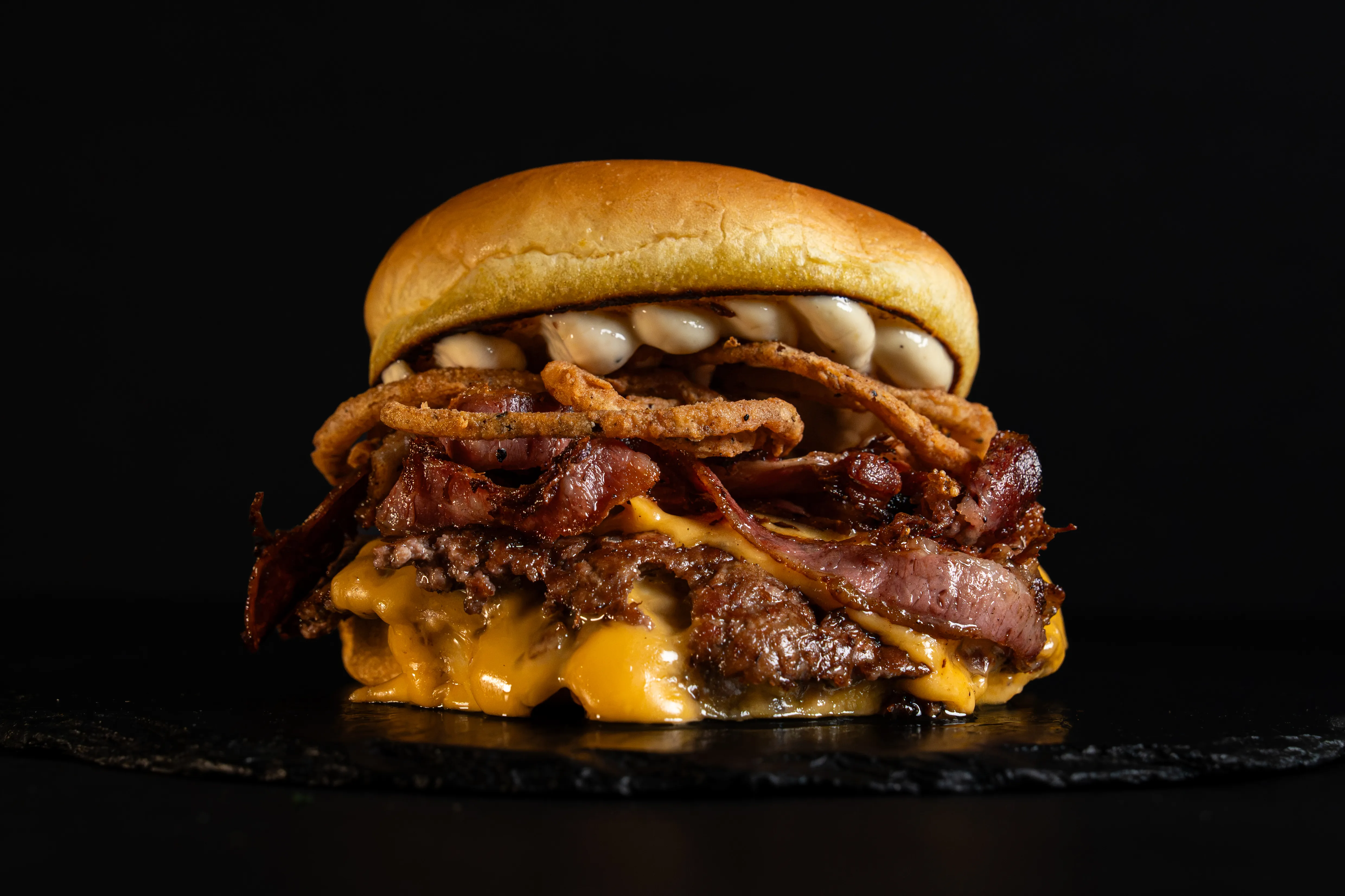 A stacked beef burger with cheese, beef bacon and crisp onion rings on a dark slate