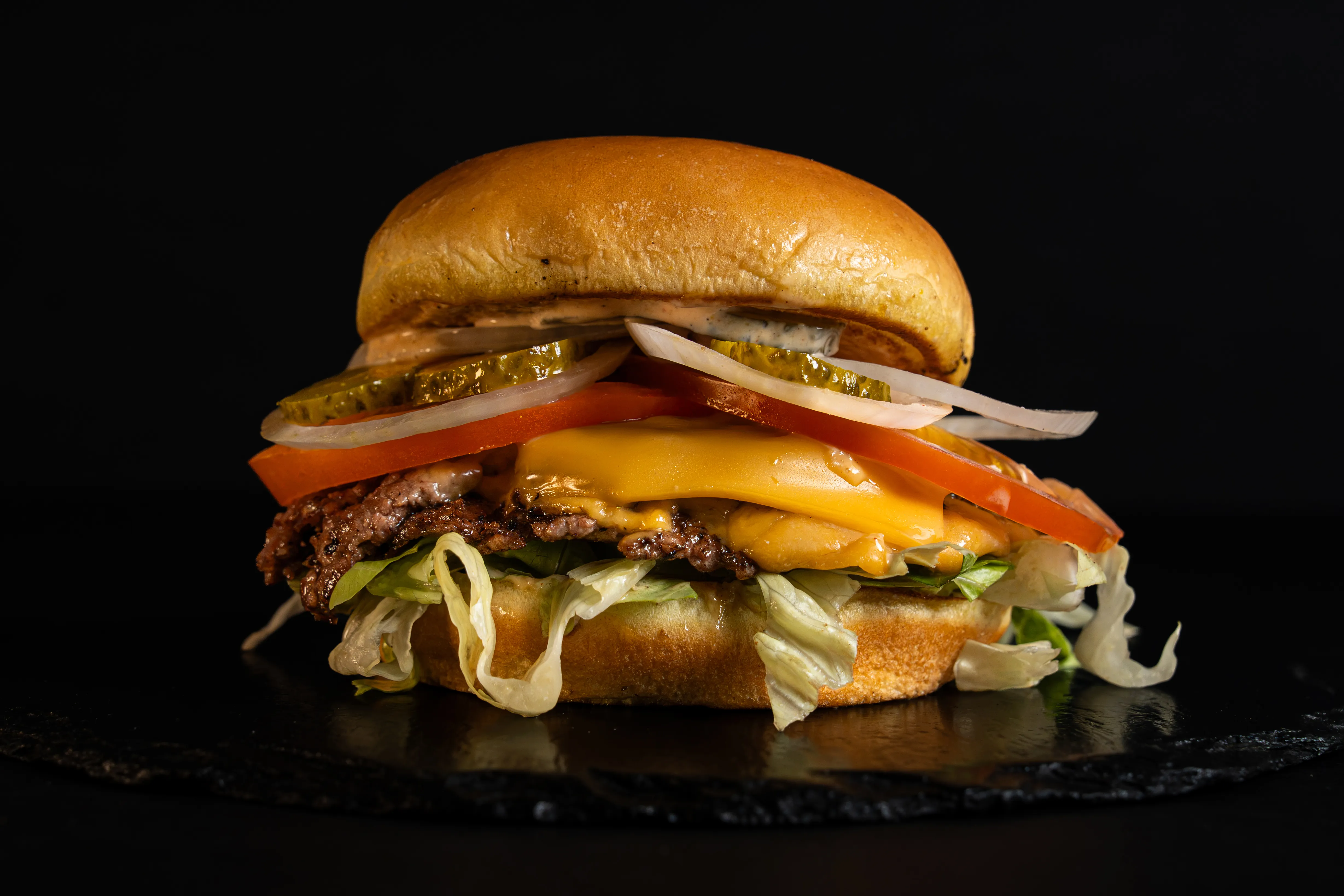 A cheeseburger with lettuce, tomato and onion on dark slate