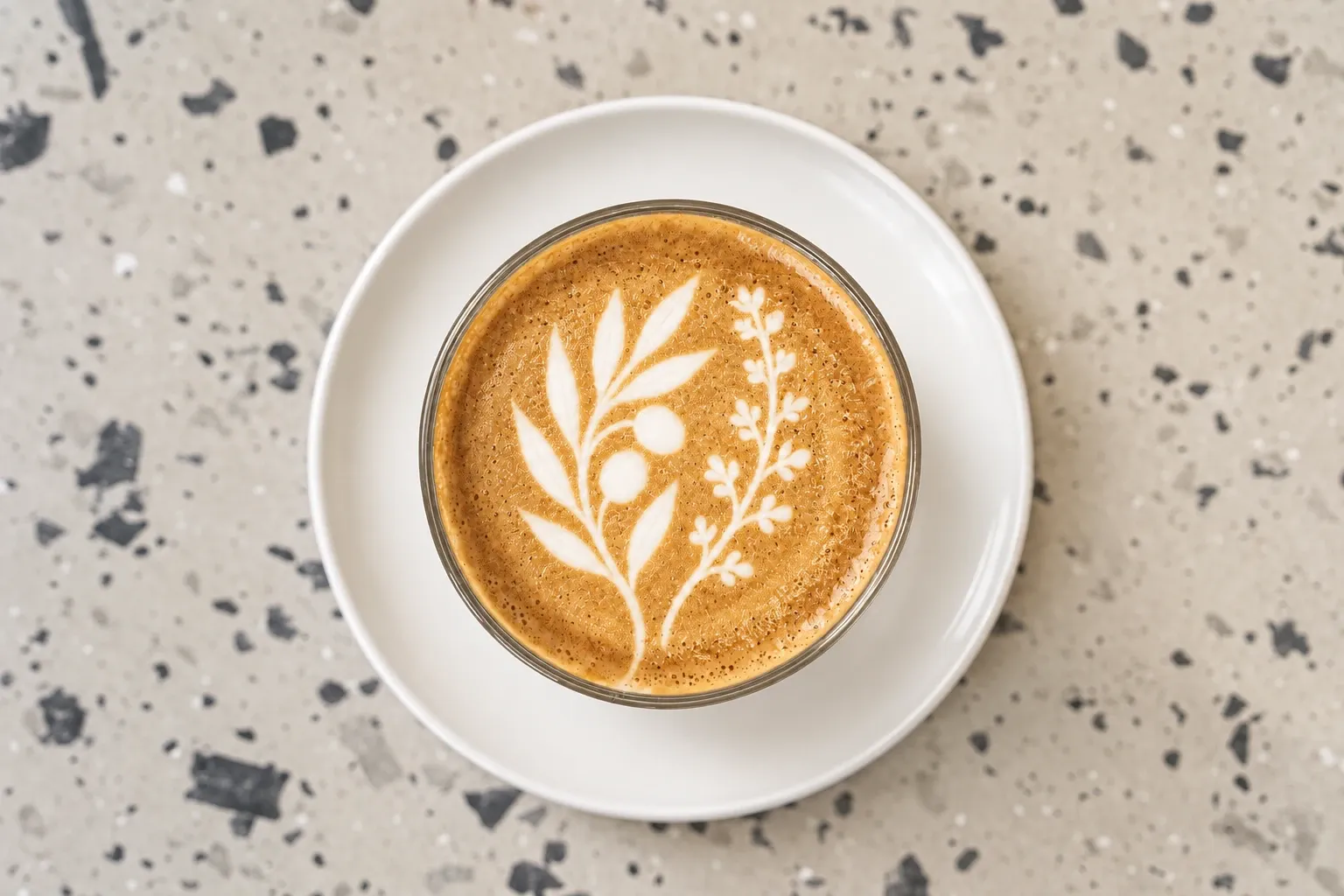 Coffee viewed from above with olive and thyme branches in the milk foam, served in a clear glass on a white saucer and pale terrazzo table