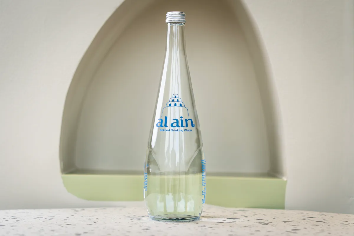 Tall clear glass Al Ain water bottle with a silver screw cap, standing in front of a beige arched wall niche with a pale green ledge